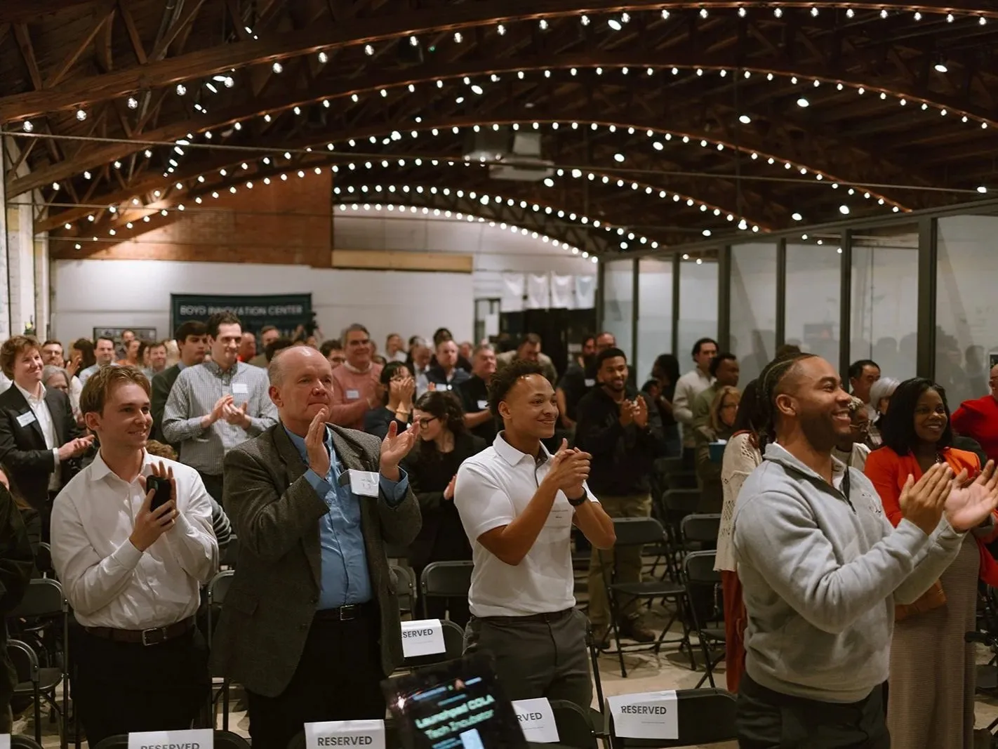 Diverse crowd applauding at a Boyd Innovation Center startup pitch night event in Columbia, SC, under string lights in a loft-style coworking space